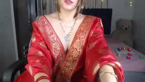 Media: Video of a fair-skinned woman in a red, gold-patterned sari, wearing a silver necklace, posing indoors. Background includes a bed with stuffed toys and colorful makeup on a table.