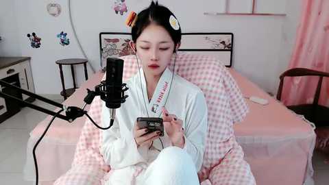 Media: Video of an East Asian woman with long black hair, wearing traditional Japanese attire, seated on a bed with pink checkered sheets, holding a smartphone, recording with a microphone.