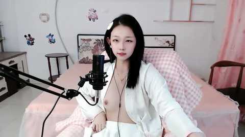 Media: Video of an East Asian woman with long black hair, wearing a white robe, sitting on a bed with pink and white bedding. She's using a microphone, recording herself.