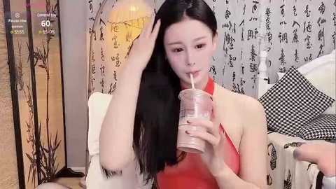 Media: Video of a fair-skinned Asian woman with long black hair, wearing a red sleeveless dress, drinking from a pink straw, in a modern living room with grey and black patterned wallpaper.