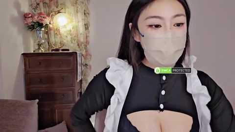 Media: Video of an Asian woman with long black hair, wearing a revealing black maid outfit with a white apron, face mask, and a \"NSFW\" watermark. Background features a wooden dresser and floral wallpaper.