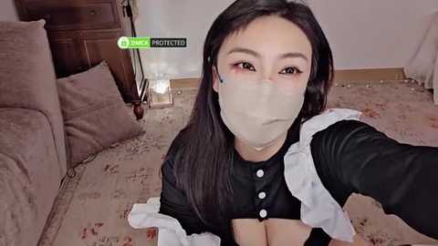 Media: A video of an Asian woman with long black hair, wearing a black maid outfit with white frills and a face mask, sitting on a beige carpet in a dimly lit room with a wooden dresser and a bed.