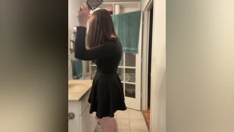 Media: Video of a woman with long brown hair, wearing a black, long-sleeved dress and a black skirt, standing in a bathroom, using a hairbrush.