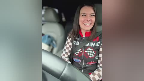 Media: Video of a smiling woman with shoulder-length dark hair, wearing a checkered racing jacket, sitting in a car with a checkered seat cover, blurred background.