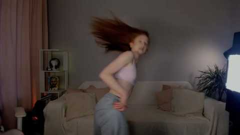 Media: A video of a young woman with long red hair, wearing a pink bra and jeans, captured in mid-motion, standing in a dimly lit living room with beige furniture and a bookshelf.