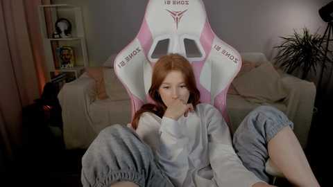 Media: Video of a young woman with fair skin and long red hair, sitting in a pink gaming chair, wearing a gray hoodie and loose pants, in a cozy living room with beige couches and bookshelves.