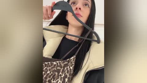 Media: Video of a woman with long black hair, wearing a beige puffer jacket, holding a leopard-print bag. She has a neutral expression. The background is blurred.