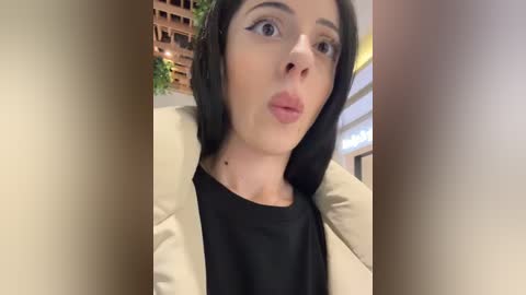 Media: Video of a fair-skinned woman with shoulder-length black hair, wearing a beige blazer over a black top, making a pouty face with exaggerated eyeliner. Background shows a modern, indoor setting with wooden beams and glass windows.