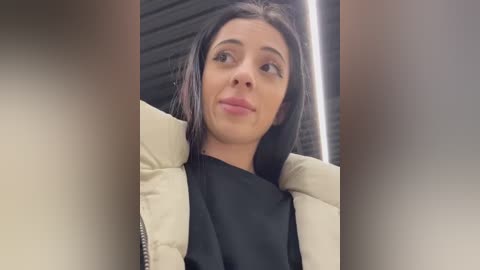 Media: Video of a young woman with fair skin, dark hair, and a confident expression, wearing a beige jacket over a black top. Background features vertical black slats.