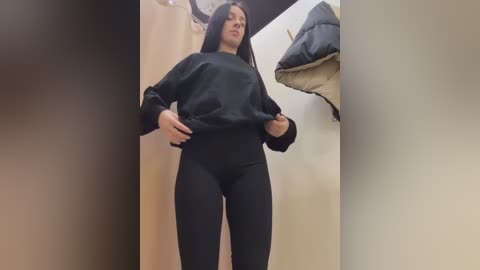 Media: Video of a young woman with long black hair, wearing a black sweatshirt and tight black leggings, standing in a dimly lit, narrow hallway.