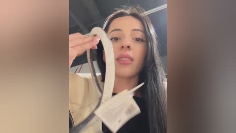 Media: Video of a young woman with long black hair, light brown skin, and full lips, holding a white envelope with a red ribbon, inside a modern room with industrial lighting.