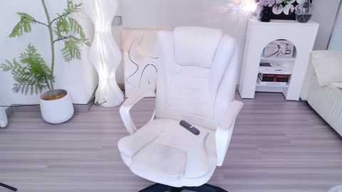 Media: Video of a modern, white ergonomic office chair with a remote control on a light wooden floor. Background features a white wall, a white bookshelf, a fern plant, and a white couch.