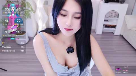 Media: A video of an East Asian woman with long black hair and light skin, wearing a low-cut, gray top, standing in a modern, white living room.