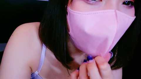 Media: Video of an Asian woman with straight black hair, wearing a light blue bra and a pink face mask, holding a blue object close to her mouth, blurred background.