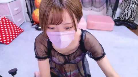 Media: Video of an Asian woman in a sheer black lingerie top, wearing a face mask, kneeling on a light carpeted floor with pink and red polka-dotted cushions. Background includes black and white decorative objects.