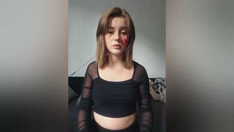 Media: A video of a young woman with shoulder-length light brown hair, wearing a black crop top and sheer sleeves, sitting indoors. She has a red streak on her forehead and a sad expression.