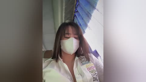 Media: A video of a woman with shoulder-length black hair, wearing a white surgical mask, and a white shirt with colorful text, standing indoors near a window with blue and white curtains.