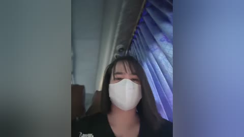 Media: Video of an Asian woman with long black hair, wearing a white surgical mask, standing indoors near a blue-lit tanning bed.