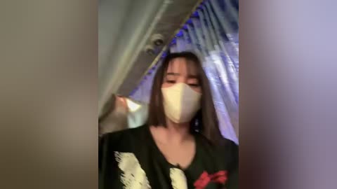 Media: Video of a person with medium-length dark hair, wearing a black shirt and white face mask, standing under a large, shiny, silver-colored object with a blue patterned background.