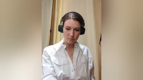 Media: Video of a woman with dark hair, wearing white button-up shirt, black over-ear headphones, and headphones around her neck, standing in a beige-walled hallway, looking down.