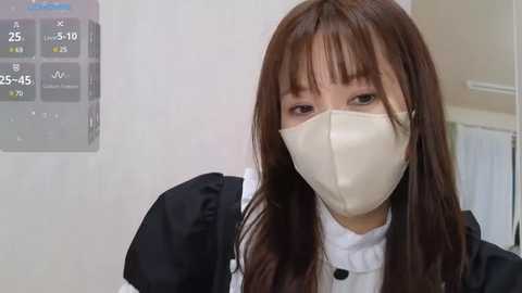 Media: Video of a young Asian woman with long brown hair and fair skin wearing a beige face mask and black and white maid outfit, standing against a white wall with a digital clock showing 25:15.