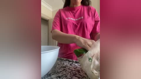 Media: Video of a woman with long brown hair, wearing a pink t-shirt, mixing green powder in a white bowl on a marble countertop.