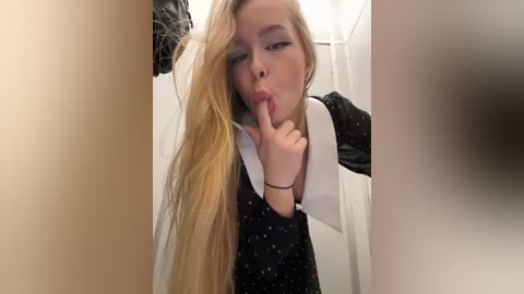 Media: Video of a young woman with long blonde hair, wearing a black polka-dot dress, making a shushing gesture in a bathroom with white walls and a shower curtain.