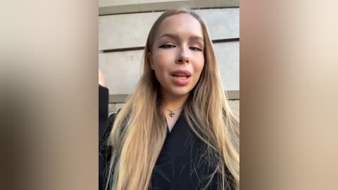 Media: Video of a young woman with long, straight blonde hair, wearing a black top, standing against a beige concrete wall. She has light skin, minimal makeup, and a neutral expression.