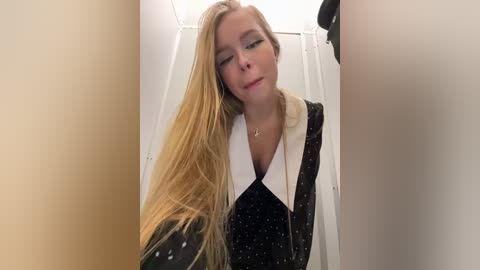 Media: Video of a young woman with long blonde hair, wearing a black dress with white collar, standing in a narrow, beige-walled hallway.