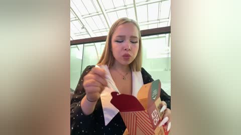 Media: Video of a blonde woman with fair skin and medium build, wearing a black and white polka dot blouse, eating a red heart-shaped lollipop and holding a red and white striped box. Background shows a modern glass building with a white ceiling.