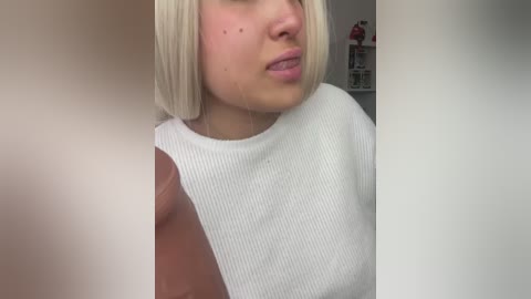 Media: Video of a fair-skinned, blonde woman with acne on her cheeks, wearing a white ribbed top. Background includes a blurred indoor scene with shelves.