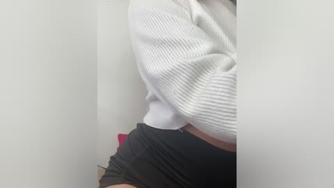 Media: A video of a woman's torso in a white ribbed sweater and black shorts, with a hint of a red object in the background, taken in a minimalist setting.