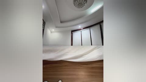 Media: A video of a modern, minimalist bedroom with white walls, a wooden floor, and a large bed covered in white sheets. The room features a circular chandelier and large windows with dark frames, creating a serene and elegant atmosphere.