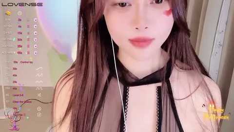 Media: Video of a young, fair-skinned woman with long brown hair, wearing a black mesh top and headphones, with a heart sticker on her cheek. Background includes a calendar and a blurred figure.