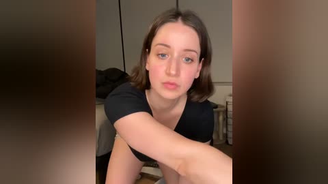 Media: Video of a young woman with fair skin, blue eyes, and straight brown hair, wearing a black top, sitting on a wooden floor in a dimly lit room.