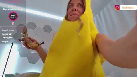 Media: A video of a blonde woman in a bright yellow jumpsuit holding a sandwich, taken from a low angle, in a modern, white-tiled room with hexagonal patterns on the wall.