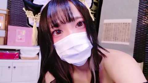 Media: Video of an Asian woman with long black hair, wearing a white surgical mask, black cat ears, and a gold headband, in a room with light-colored walls and a white cabinet.