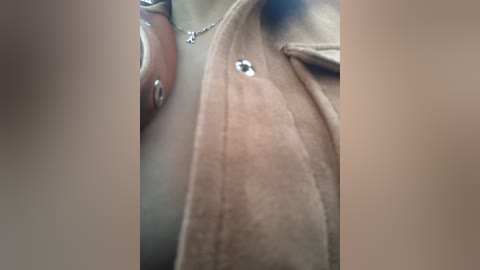 Media: Video of a person's hand holding a light brown suede jacket with silver buttons and a delicate silver necklace. The background is blurred, focusing on the details of the clothing.