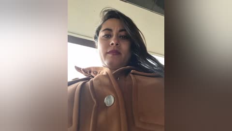 Media: A close-up video of a woman with medium brown skin, dark hair, and a neutral expression, wearing a brown coat, captured from a low angle.