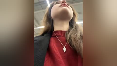 Media: Video of a young woman with fair skin, wearing a red turtleneck sweater and a black blazer, with long brown hair. She has a silver necklace with a heart pendant. Background shows an indoor setting with fluorescent lighting.