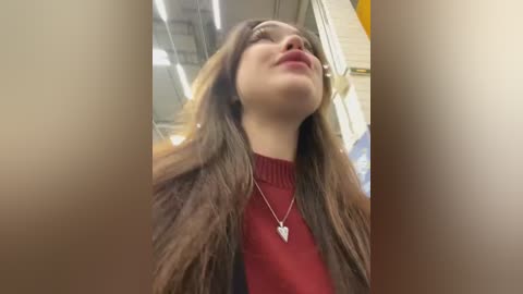 Media: A video of a young woman with long brown hair, fair skin, and red lipstick, wearing a red turtleneck sweater, standing in an indoor, bright, modern setting with fluorescent lights and a yellow wall in the background.