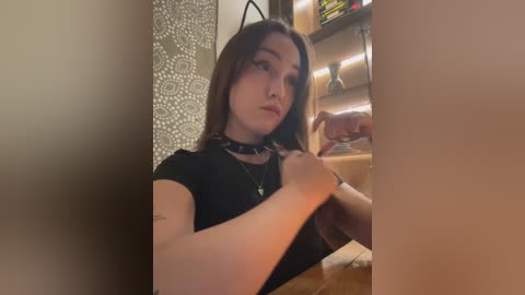 Media: Video of a young woman with long brown hair, wearing a black choker and top, adjusting a necklace in a dimly lit room with a patterned wall and wooden shelves.