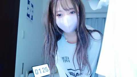 Media: A video of a young Asian woman with long, straight brown hair, wearing a white mask and a white shirt with black text, standing in a room with a digital clock reading \"02:25.\