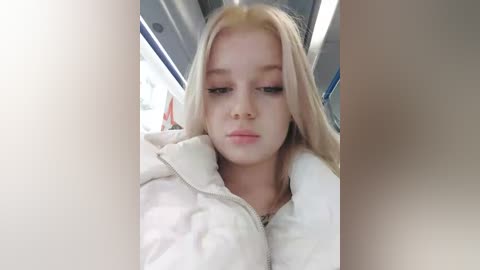 Media: Video of a young, fair-skinned girl with long, blonde hair, wearing a white puffy jacket, seated on a bus. She has a neutral expression, surrounded by blurred, beige bus windows.