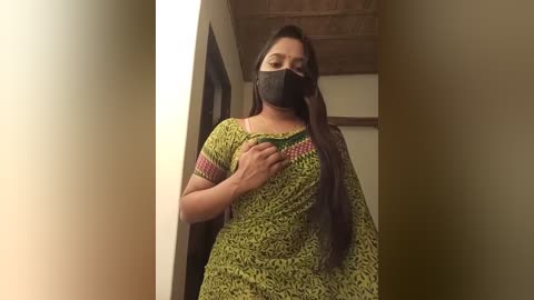 Media: Video of a South Asian woman with long dark hair, wearing a green dress with a patterned design, a black face mask, and a black headband. She stands in a narrow, dimly lit hallway with beige walls and a wooden ceiling.