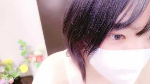 Media: Video of a young Asian woman with shoulder-length black hair, wearing a white face mask, in a softly focused indoor setting with blurred floral arrangements in the background.