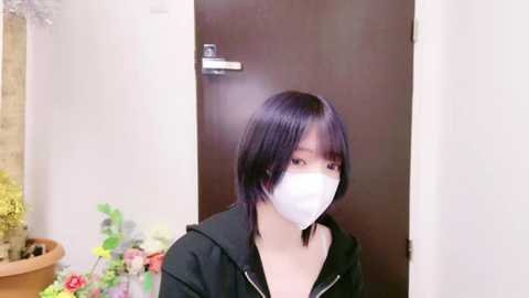 Media: Video of an Asian woman with short black hair, wearing a white face mask, black hoodie, and standing in a room with a potted plant and a brown door.