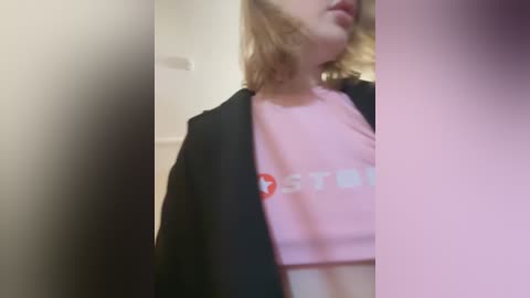 Media: A video of a young woman with shoulder-length, wavy blonde hair, wearing a pink crop top with \"STY\" in white letters, and a black blazer, partially blurred, in a neutral-toned room.