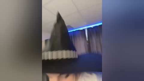 Media: Video of a person wearing a black witch hat and cloak, blurred, standing in a dimly lit room with blue LED strip lighting and beige curtains.