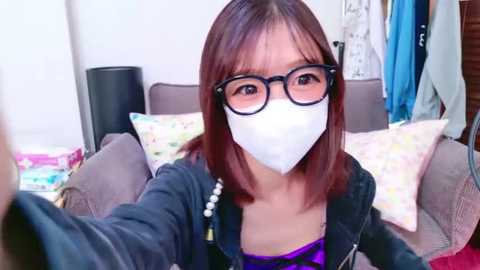 Media: Video of an Asian woman with shoulder-length brown hair, wearing a face mask, glasses, and a black jacket, taking a selfie in a cozy living room with patterned pillows and a white curtain.